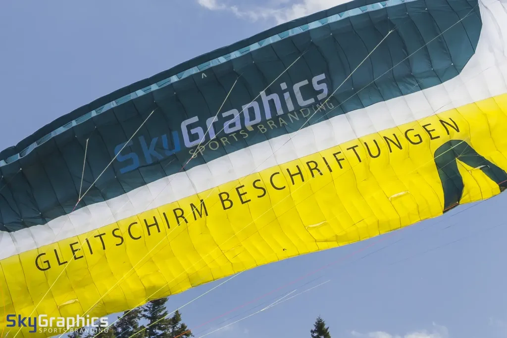 Beschrifteter Gleitschirm im Flug mit großem Schriftzug auf gelbem und petrolfarbenem Tuch